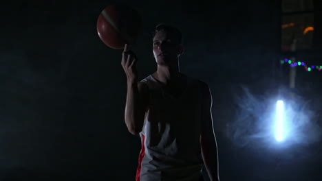 Un-Hombre-En-Forma-Deportiva-Gira-Una-Pelota-De-Baloncesto-En-Su-Dedo-Realizando-Un-Truco-Mirando-A-La-Cámara-En-La-Cancha-De-Baloncesto-Alrededor-Del-Humo-En-Cámara-Lenta.